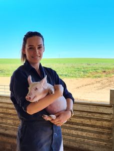 a young woman dressed in a dark blue coverall holding a piglet on a farm